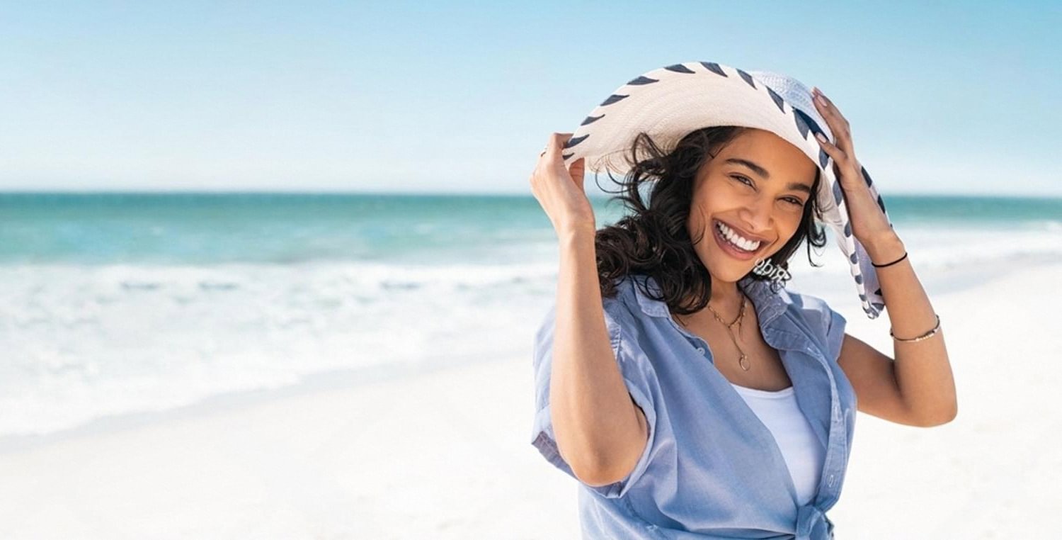Woman in sun hat smiling on a UAE beach practicing summer skin protection